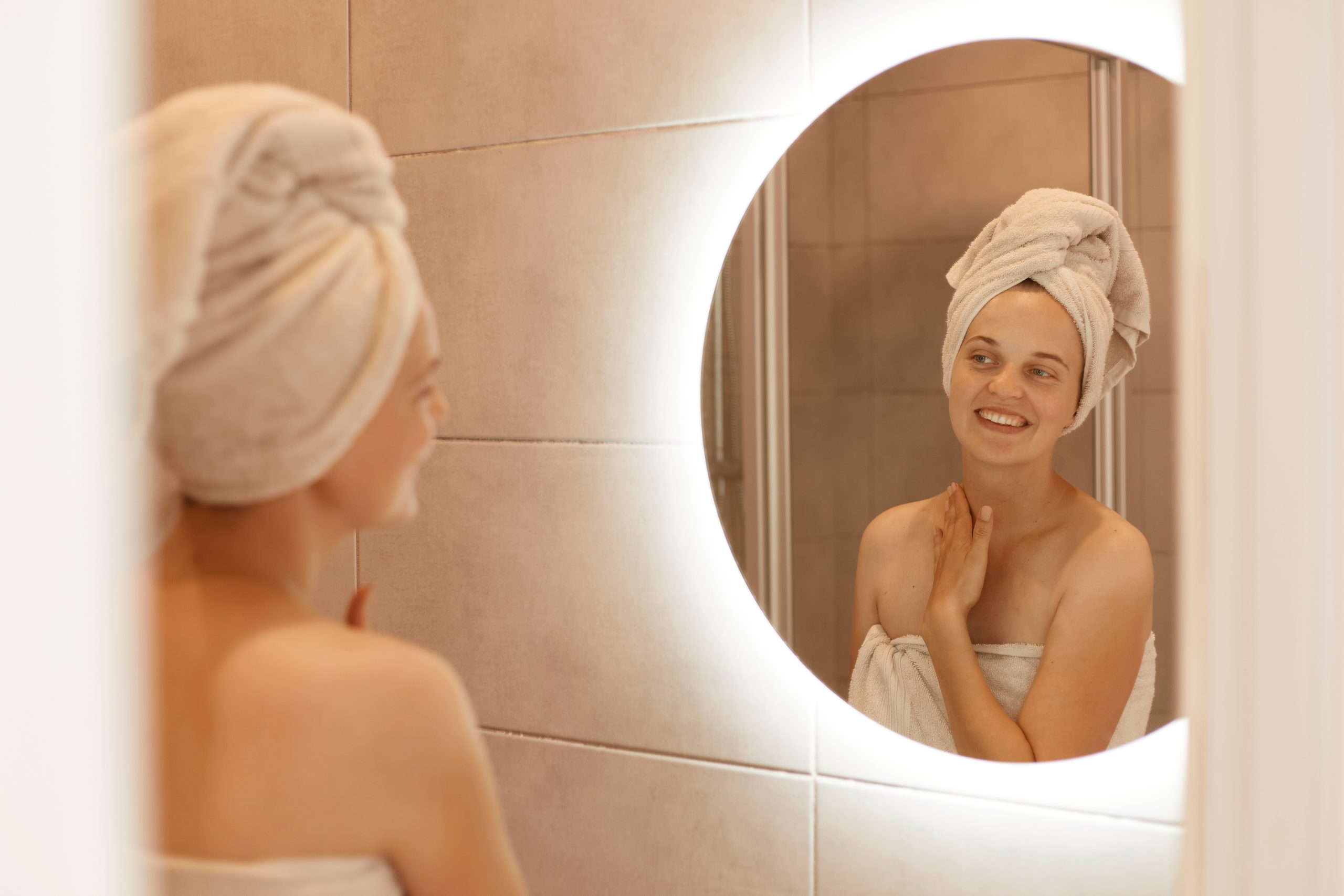 Portrait of beautiful Caucasian woman with towel on her hair touching her neck at bathroom, looking at her reflection in the mirror, posing after taking shower.