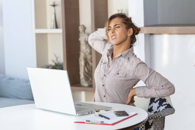 portrait young stressed woman sitting home office desk front laptop touching aching back with pained expression suffering from backache after working laptop wp